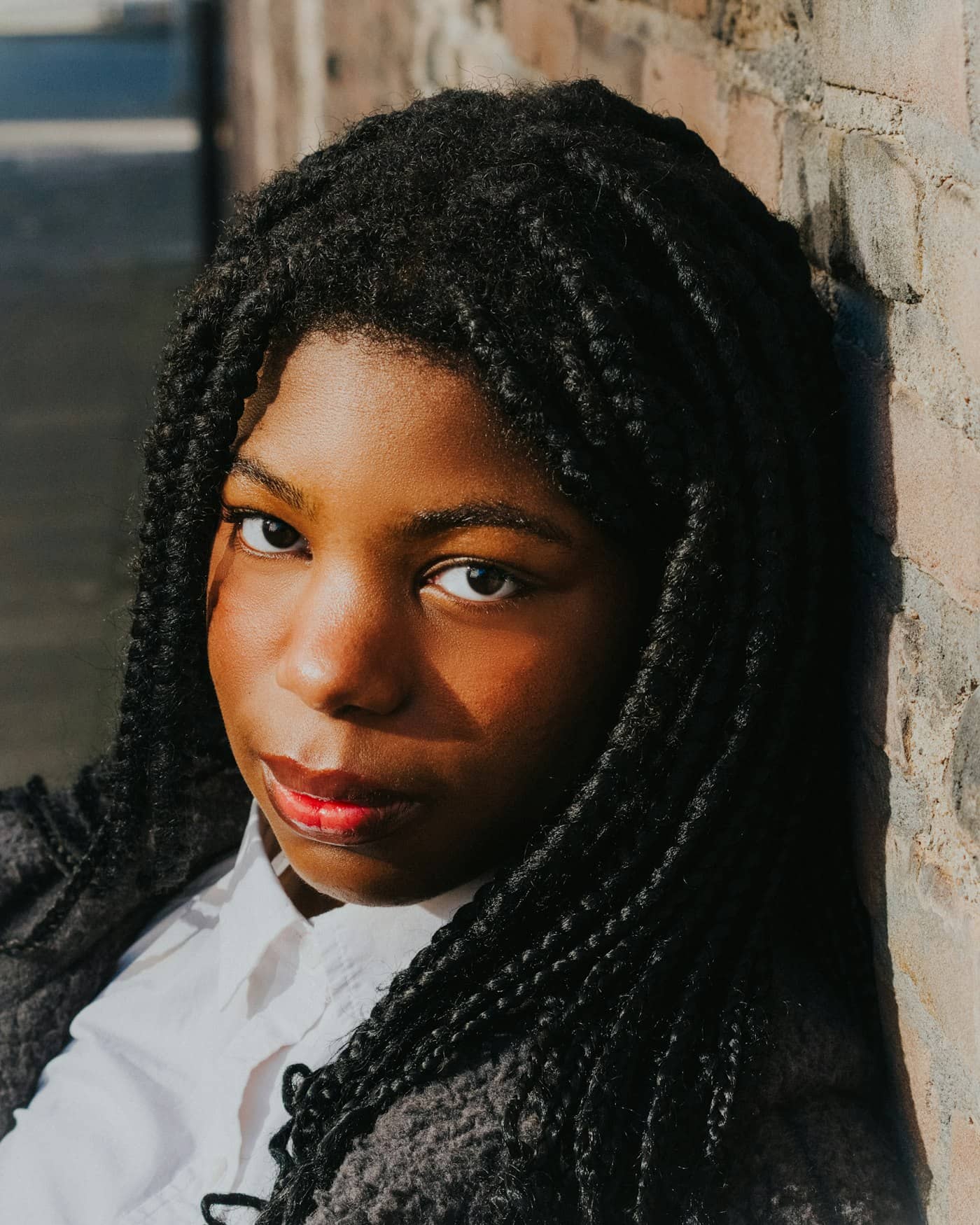 Portrait d'une jeune femme aux box braids volumineuses, adossée à un mur de briques